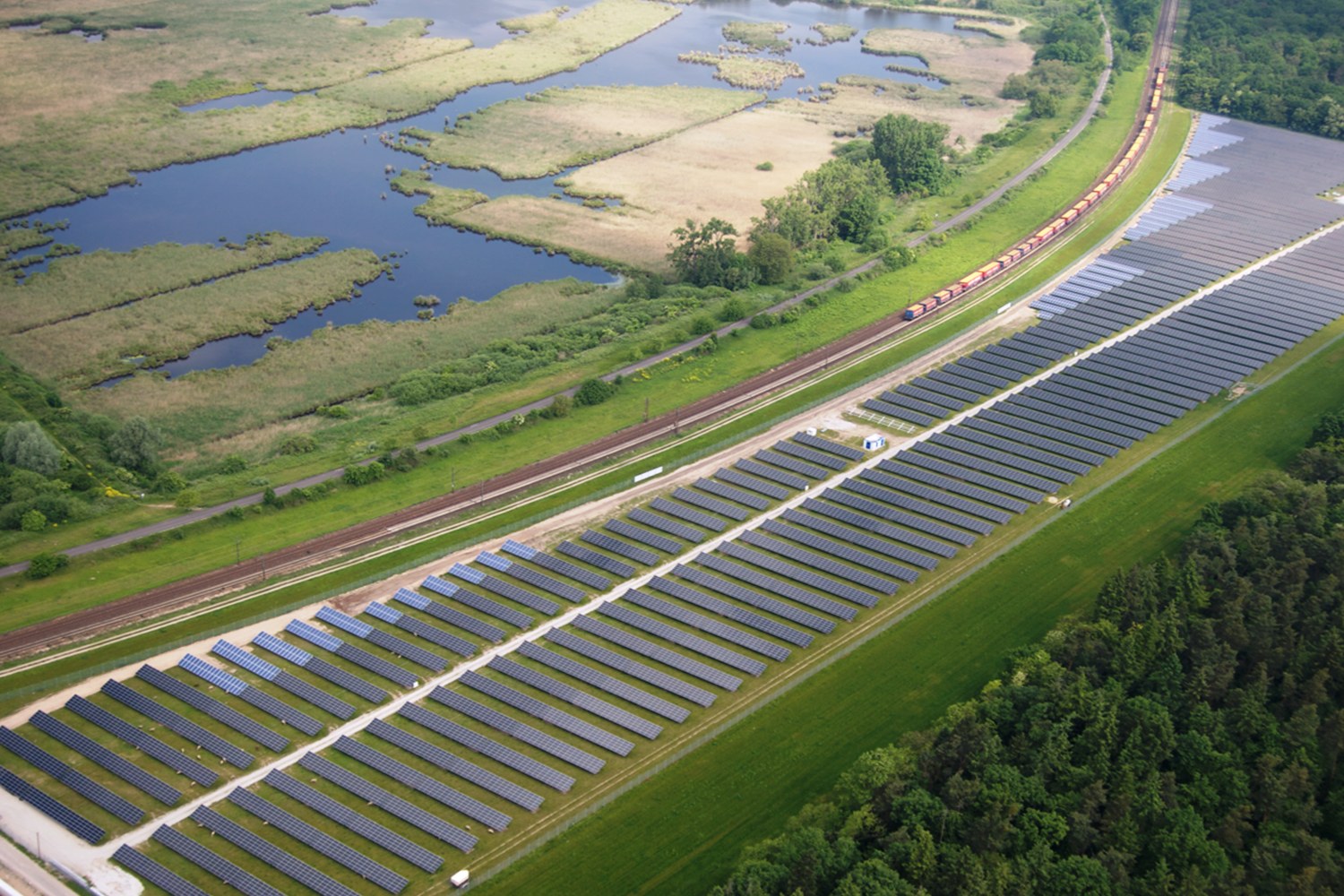 Luftaufnahme eines Solarparks mit Solarpanelen, umgeben von Bäumen, Gras, Pflanzen und Wasser, mit einem Zug auf einer nahen Eisenbahnstrecke.