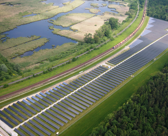 Luftaufnahme eines Solarparks mit Solarpanelen, umgeben von Bäumen, Gras, Pflanzen und Wasser, mit einem Zug auf einer nahen Eisenbahnstrecke.