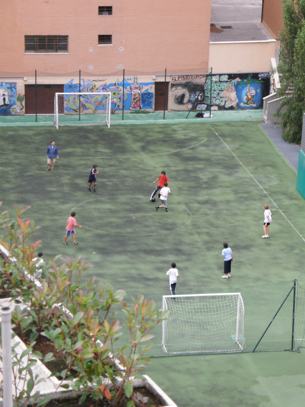 Kinder spielen Fußball auf einem Feld mit Toren, ein Gebäude mit Graffiti im Hintergrund, Pflanzen links und Bäume rechts.