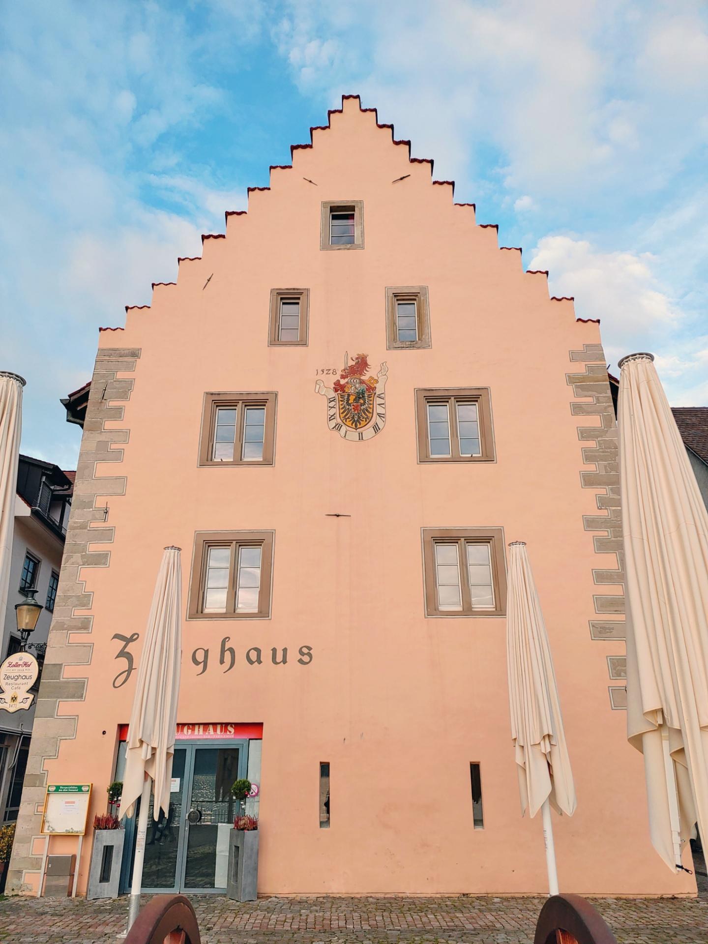 Pinkes Gebäude mit einem zentralen Uhrenturm, umgeben von Fenstern, Regenschirmen, einem Schild, einer Straßenlaterne, Topfpflanzen und einem bewölkten Himmel, repräsentiert München, Deutschland.