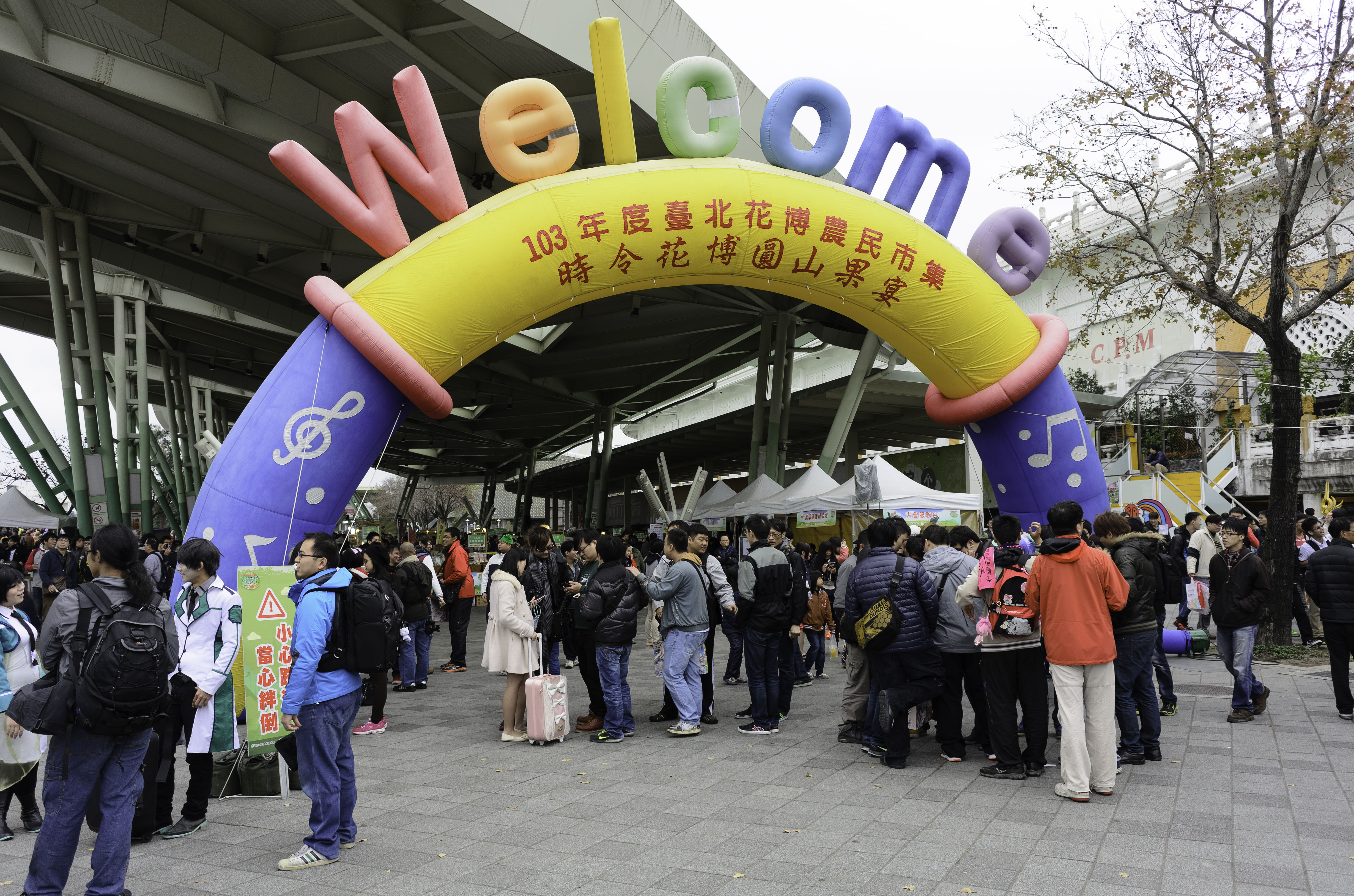 Eine Gruppe von Menschen steht vor einem Willkommensbogen mit Text, umgeben von Zelten, Bäumen, Gebäuden und einem klaren blauen Himmel, einige tragen Taschen.