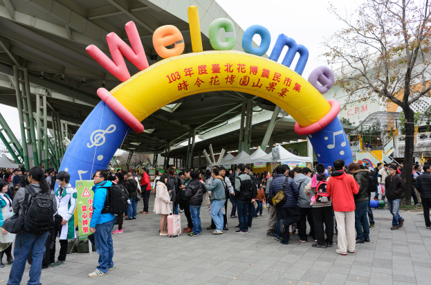 Eine Gruppe von Menschen steht vor einem Willkommensbogen mit Text, umgeben von Zelten, Bäumen, Gebäuden und einem klaren blauen Himmel, einige tragen Taschen.