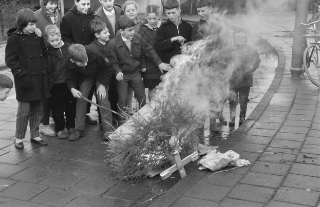 Schwarzes Bild von Kindern, die sich um ein Straßenfeuer versammeln, einige halten Stöcke, mit Fahrrädern, Pfählen, Bäumen und Gebäuden im Hintergrund.