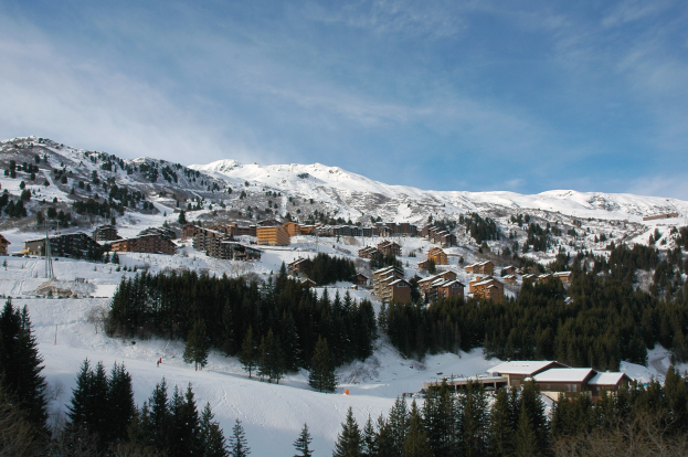Ein malerischer Blick auf ein Skigebiet in den französischen Alpen mit schneebedeckten Bergen, Bäumen, Gebäuden und einem klaren blauen Himmel.