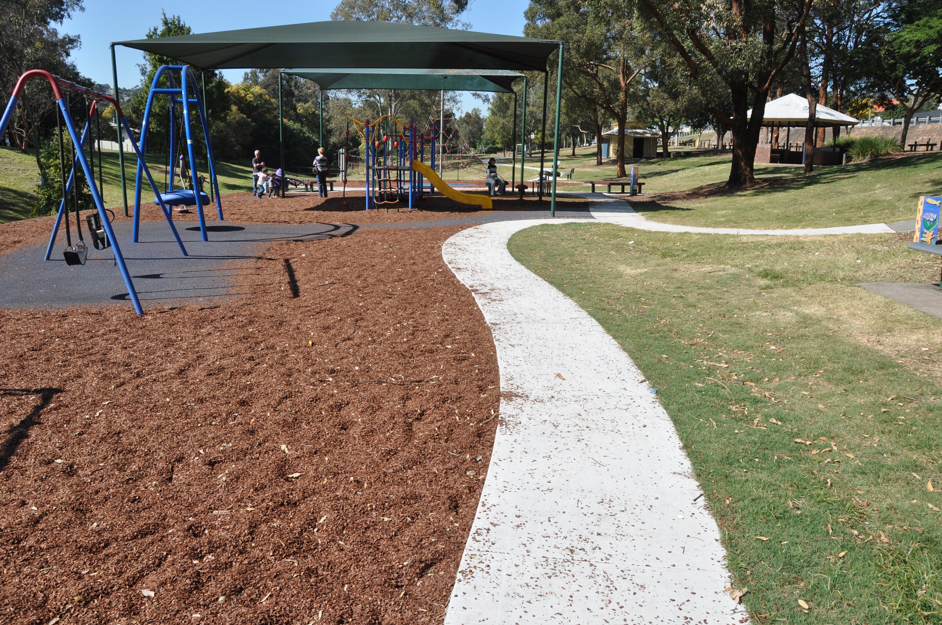 Ein Park mit einem Spielplatz, der über Schaukeln und Rutschen verfügt und von grünem Gras, Bäumen, Bänken, Hütten im Hintergrund und einem klaren blauen Himmel umgeben ist.