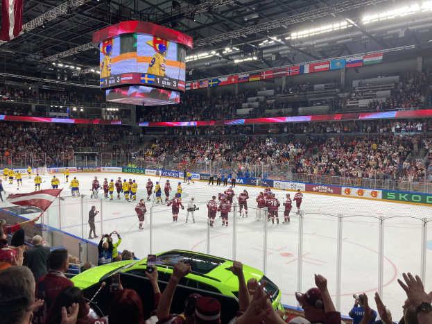 Ein Eishockeyspiel wird in einer großen Arena mit Zuschauern gespielt, einige halten Fahnen, und ein Auto steht davor. Die Eisfläche ist von einem Zaun umgeben und Banner mit Text, während die Decke mit Lichtern, einem Bildschirm und hängenden Fahnen geschmückt ist.