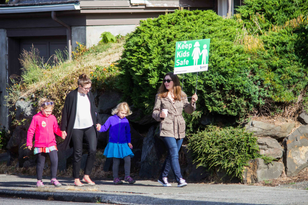 Eine Gruppe von Menschen geht eine Straße entlang, mit einer Frau in Fürhrung, die ein Schild mit der Aufschrift "Kinder schützen" hält, Straße gesäumt von Bäumen, Pflanzen und Felsen, mit einem Gebäude im Hintergrund.