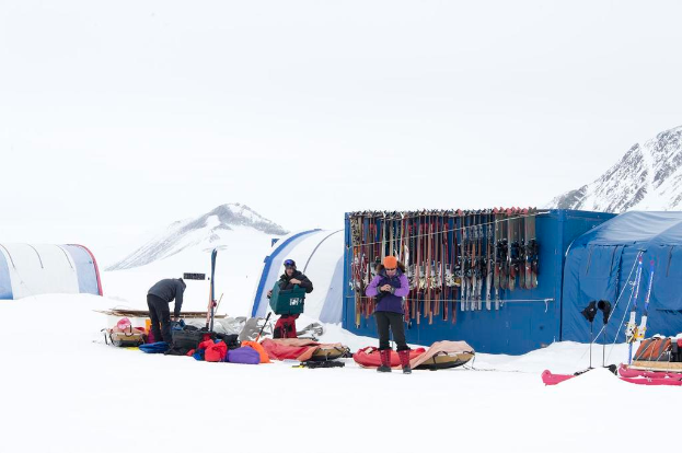 Drei Personen stehen auf einer schneebedeckten Landschaft mit Ã¼berall verstreuten Taschen, Zelten mit Skiern darauf und schneebedeckten HÃ¤ngen im Hintergrund unter einem klaren Himmel.