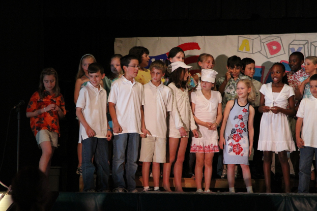 Mehrere Kinder auf einer Bühne, wobei zwei in der Mitte mit Kappen stehen, ein Mikrofonständer links und ein Banner im Hintergrund.