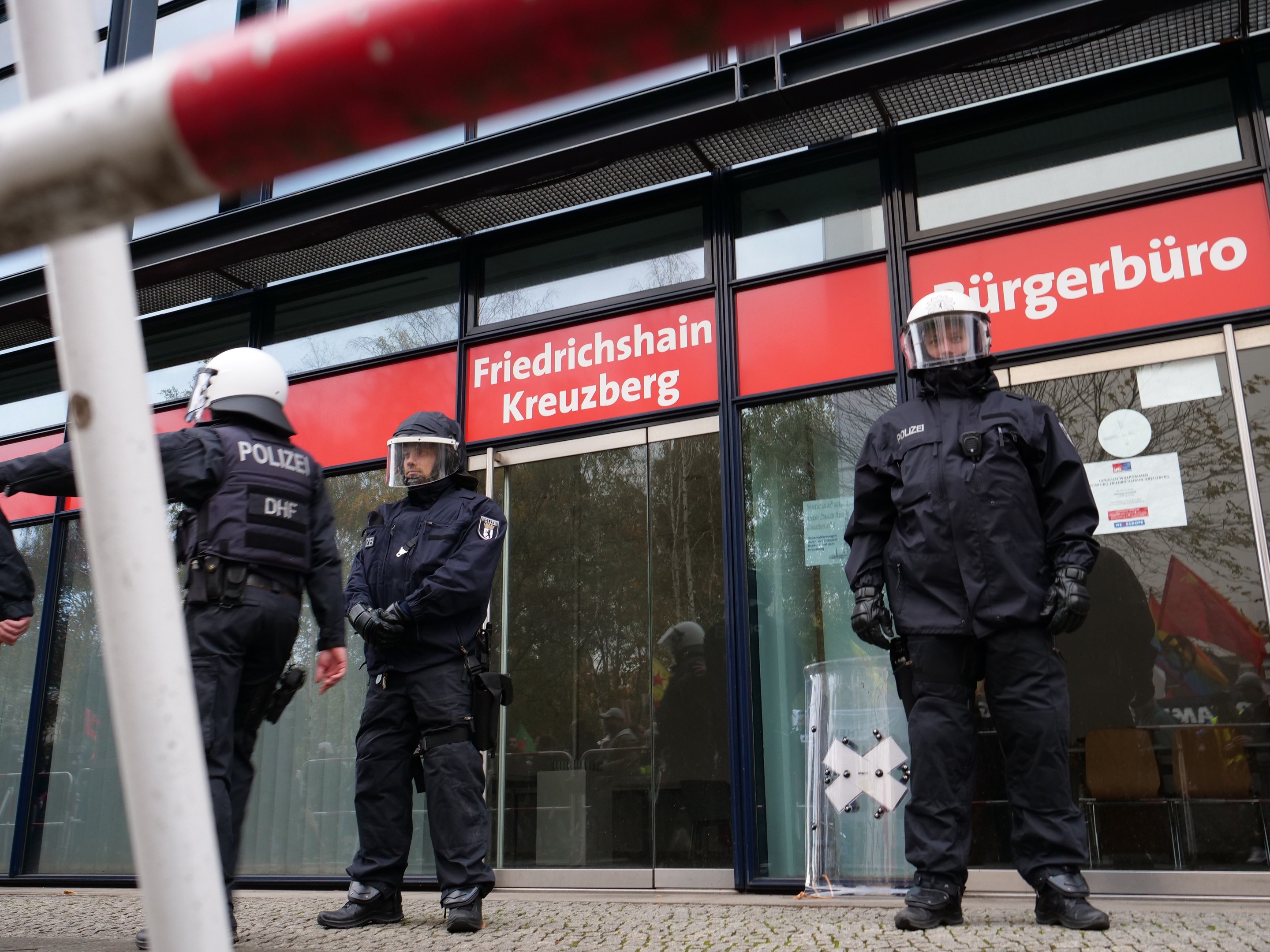 Polizisten in Uniform stehen vor einem Gebäude mit Glaswänden und -türen, tragen Helme und Handschuhe und halten einen Stock in der linken Hand. Im Hintergrund sind Schilder mit der Aufschrift "Friedrichshain Kreuzberg" zu sehen.