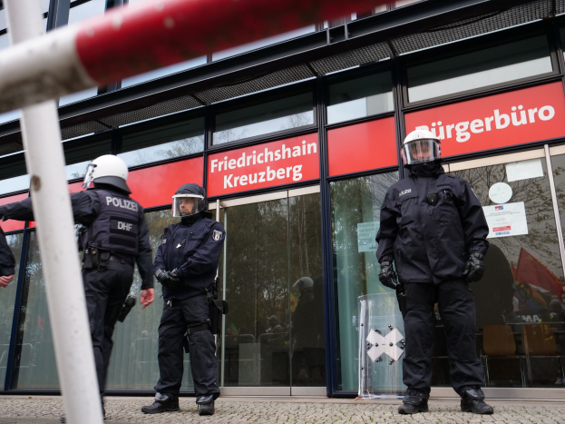 Polizisten in Uniform stehen vor einem Gebäude mit Glaswänden und -türen, tragen Helme und Handschuhe und halten einen Stock in der linken Hand. Im Hintergrund sind Schilder mit der Aufschrift "Friedrichshain Kreuzberg" zu sehen.