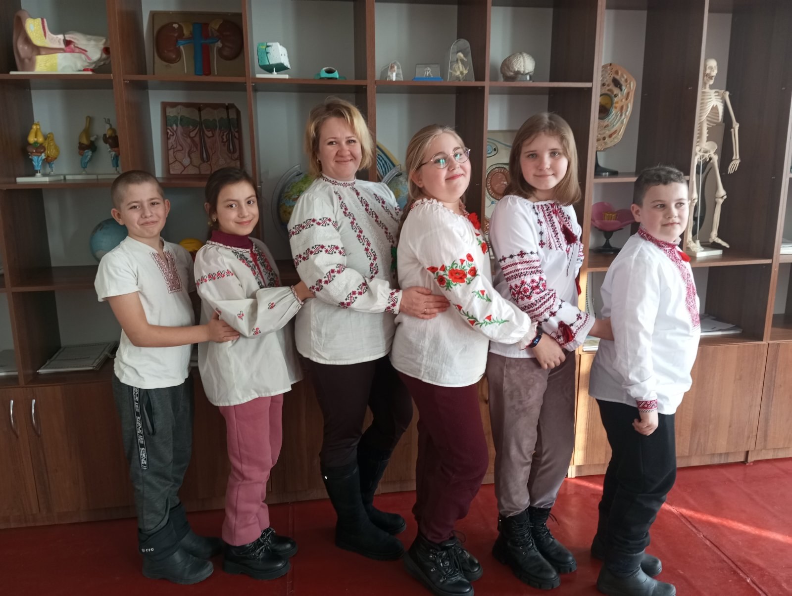 Eine Gruppe von Kindern in traditioneller ukrainischer Kleidung vor einem Bücherregal mit Spielzeug, einem Skelett und anderen Gegenständen in einer ukrainischen Schulumgebung.