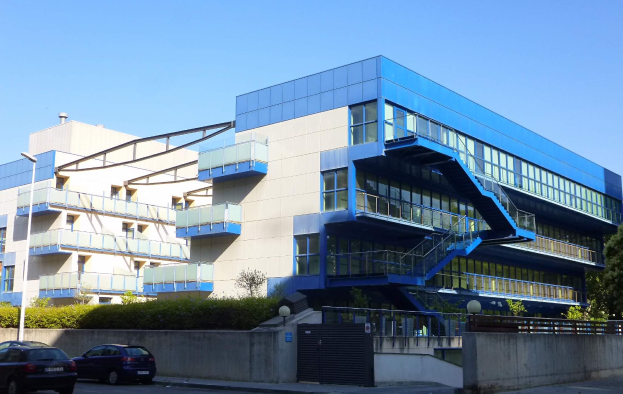 Großes modernes Gebäude mit zahlreichen parkenden Autos davor, umgeben von Straßenlaternen, Lampen, Gittern, Grünflächen und einem klaren blauen Himmel, identifiziert als der neue Hauptsitz der University of California, Berkeley.