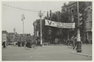 Ein Schwarz-Weiß-Foto einer Stadtstraße mit Menschen auf Fahrrädern und zu Fuß, Ampeln, Laternenpfähle, Bäume und Gebäude im Hintergrund bei klarem Himmel.