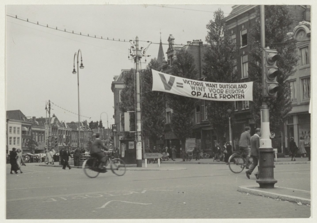 Ein Schwarz-Weiß-Foto einer Stadtstraße mit Menschen auf Fahrrädern und zu Fuß, Ampeln, Laternenpfähle, Bäume und Gebäude im Hintergrund bei klarem Himmel.