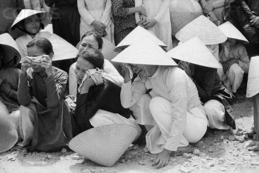 Ein Schwarz-Weiß-Bild von einer Gruppe von Menschen, die auf dem Boden sitzen, Hüte tragen und weinen, mit Steinen unten.