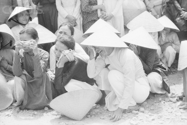 Ein Schwarz-Weiß-Bild von einer Gruppe von Menschen, die auf dem Boden sitzen, Hüte tragen und weinen, mit Steinen unten.