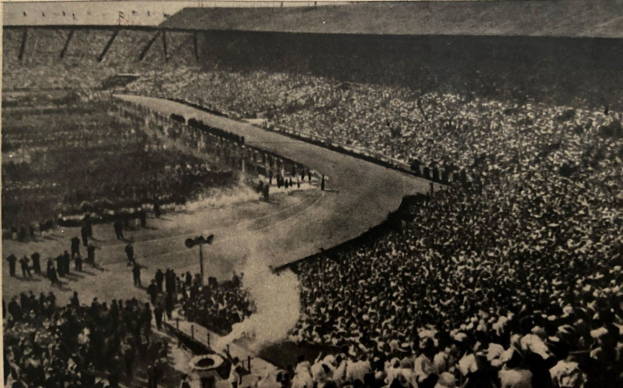 Schwarzes und weißes Foto einer großen Menschenmenge in einem Stadion mit einer zentralen Straße und Pfählen auf beiden Seiten, das Text am unteren Rand enthält.
