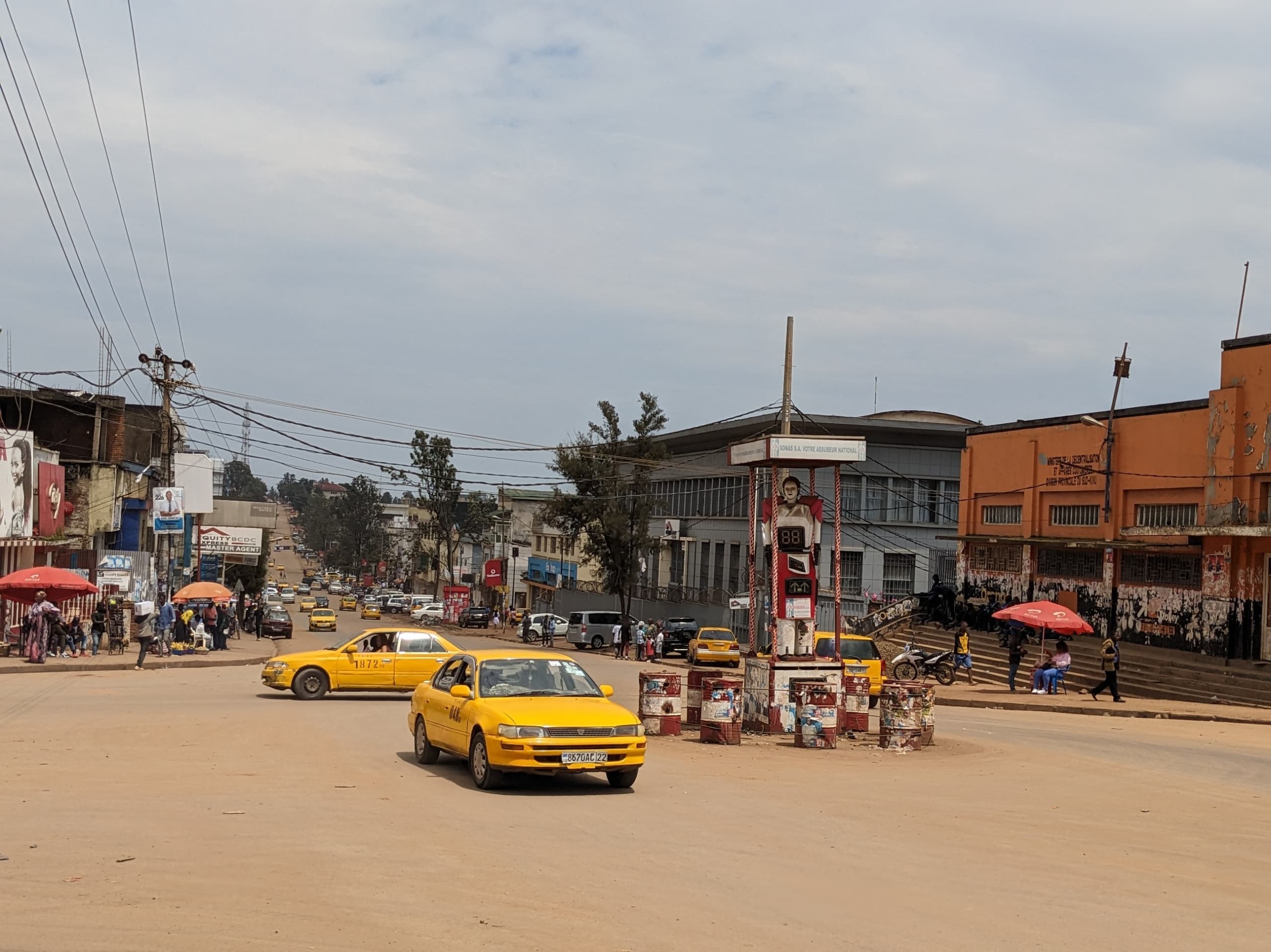 Ein gelbes Taxi fährt auf einer belebten Stadtstraße mit Gebäuden an den Seiten, während Fußgänger mit Regenschirmen auf dem Gehweg unter einem bewölkten Himmel gehen.