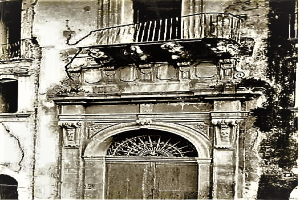 Ein altes Gebäude mit einer Tür, Fenstern, Geländern und Skulpturen an seiner Fassade, dargestellt in Schwarz-Weiß.