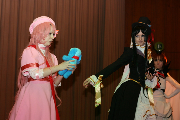 Drei Frauen sind in einer dramatischen Performance zu sehen, wobei eine in einem pinkfarbenen Kleid einen blauen Teddybär hält, eine andere in einem schwarzen Kleid nach links schaut und ein brauner Vorhang im Hintergrund zu sehen ist.