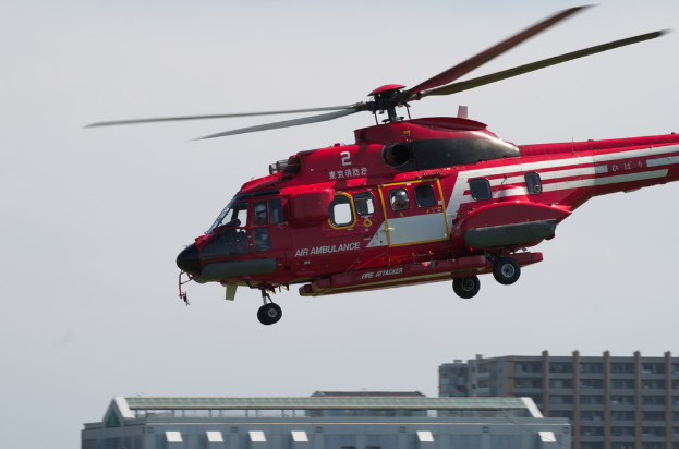 Ein roter Hubschrauber fliegt nach links mit Gebäuden unten und einem Himmel im Hintergrund.