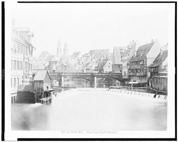 Ein Schwarz-Weiß-Foto eines Flusses mit einer Brücke darüber, umgeben von Gebäuden, Bäumen und einem Himmel, mit dem Text "Nürnberg, Blick von Karl's Brücke" unten.