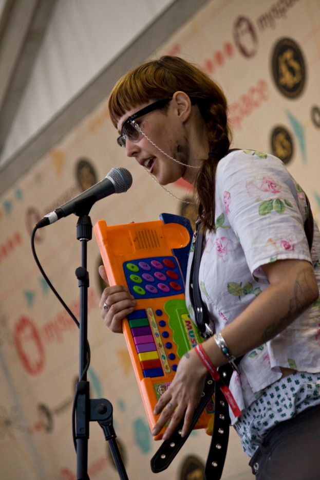 Eine Frau in einem weißen Hemd und Sonnenbrille singt in ein Mikrofon und spielt Piano, mit einem Plakat im Hintergrund.