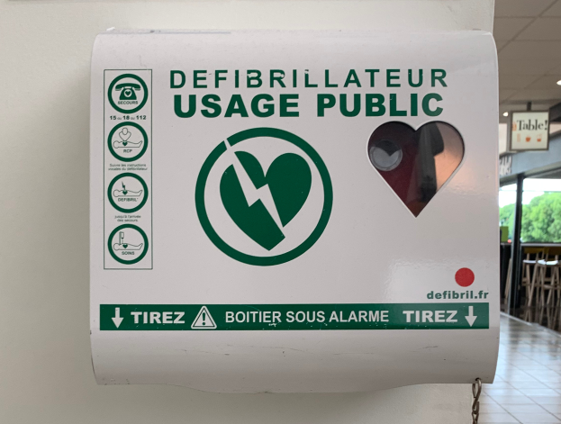 Ein öffentliches Defibrillator-Gebrauchszeichen an einer Gebäudewand, umgeben von Stühlen, Tischen, Brettern, Lampen und Bäumen im Hintergrund und der Boden unten sichtbar.