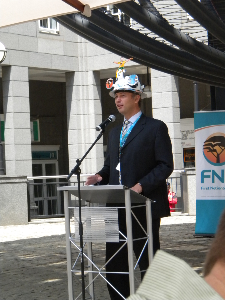 Ein Mann in Anzug und Hut spricht in ein Mikrofon an einem Rednerpult, mit einer Person rechts unten, einem Banner mit Text rechts und einem Gebäude mit Geländer im Hintergrund.