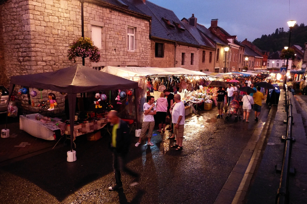 Eine Gruppe von Menschen auf der Straße, einige mit Schirmen, Geschäfte mit hängenden Spielzeugen, Taschen, Gebäude mit Fenstern, Bäume, Straßenlaternen und Barrieren.
