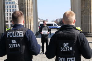 Drei Polizeibeamte stehen vor einem Gebäude, ein Mann hält in der Mitte ein Papier, flankiert von Säulen, mit Fahrzeugen, Menschen, Bäumen, Gebäuden und Himmel im Hintergrund.
