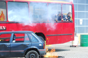 Roter Doppeldeckerbus mit Rauch, drei sichtbaren Passagieren, neben einem Auto geparkt, mit einem Glasfenster-Gebäude und einem Fass im Hintergrund.