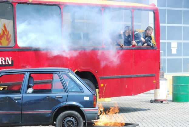 Roter Doppeldeckerbus mit Rauch, drei sichtbaren Passagieren, neben einem Auto geparkt, mit einem Glasfenster-Gebäude und einem Fass im Hintergrund.