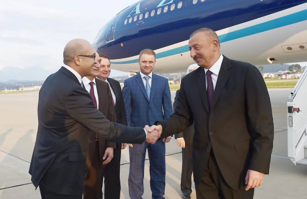 Aserbaidschanischer Präsident İlham Əliyev und türkischer Präsident Recep Tayyip Erdoğan geben sich die Hand vor einem Flugzeug, umgeben von einer Gruppe von Menschen, mit einem Fahrzeug auf der rechten Seite und Gebäuden, Bäumen, Gras, Hügeln und einem klaren blauen Himmel im Hintergrund.