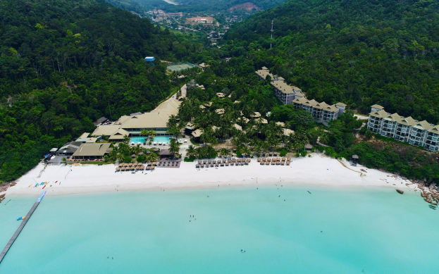 Luftaufnahme eines Resorts am Strand von Koh Samui, mit einer Brücke links, Gebäuden, Bäumen, Hügeln und Himmel im Hintergrund sowie einigen Menschen in Strandnähe.