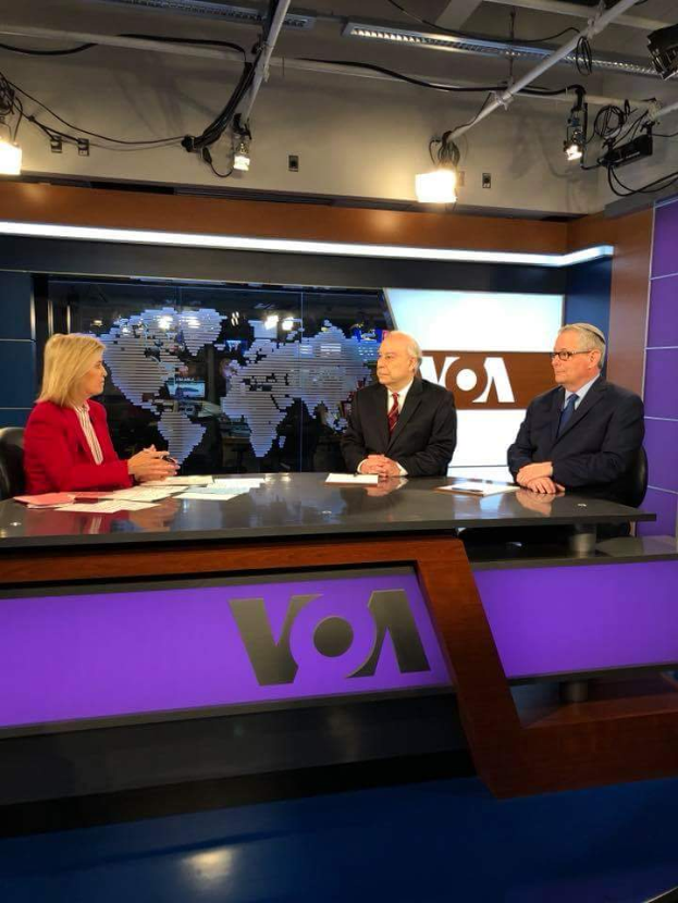 Drei Personen, zwei Männer in Blazern und Krawatten und eine Frau in einem roten Kleid, sitzen an einem Schreibtisch mit Papieren und anderen Gegenständen und führen eine Diskussion vor einem Fernseher mit einem Bildschirm und Lampen im Hintergrund.