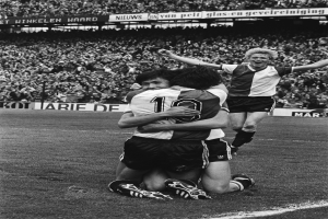 Drei Fussballspieler feiern ein Tor auf dem Boden in einem schwarz-weissen Stadion.
