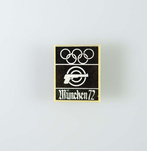 Ein schwarzes und weißes Logo mit einem schwarzen Kreis mit weißer Umrandung, der eine weiße Silhouette einer Person mit ausgebreiteten Armen enthält, sowie der Text "München 1972" in fetter schwarzer Schrift.