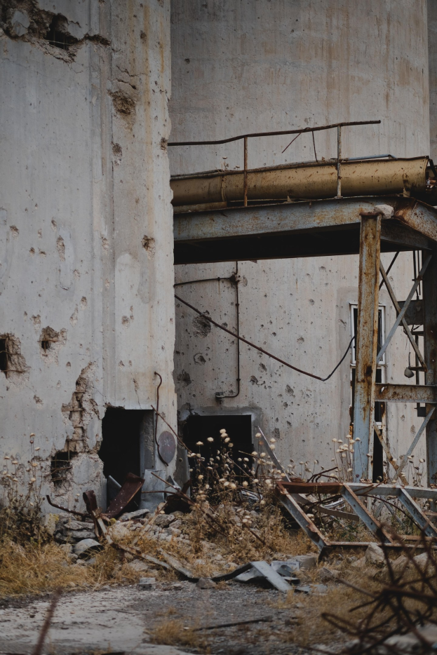 Altes Gebäude mit einem zentralen Metallgerüst, umgeben von verstreuten Pflanzen, Eisenstangen und Trümmern, mit bröckelnden Wänden und zerbrochenen Fenstern.