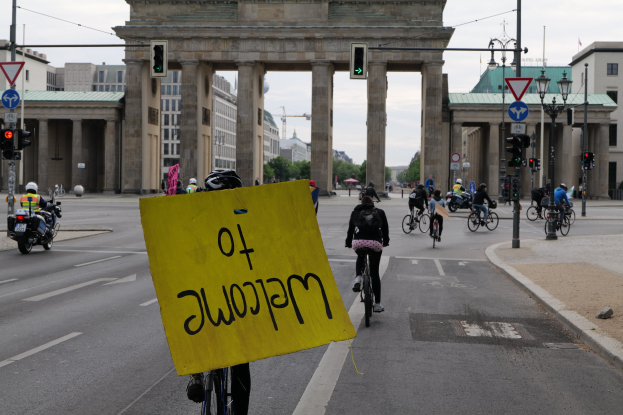 Eine Gruppe von Radfahrern mit Helmen fährt an der Brandenburgertor in Berlin vorbei, einer hält ein gelbes Schild, mit Laternenmasten, Verkehrszeichen, Gebäuden, Bäumen und einem klaren blauen Himmel im Hintergrund.