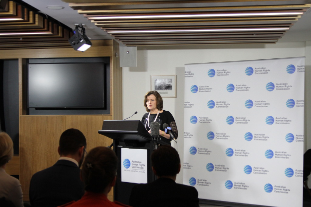 Eine Frau steht auf einer Bühne und spricht in ein Mikrofon, vor ihr sitzt ein Publikum, an der Wand hängt ein Banner, auf der hölzernen Wand steht ein Fernseher, und an der Decke sind Lampen.