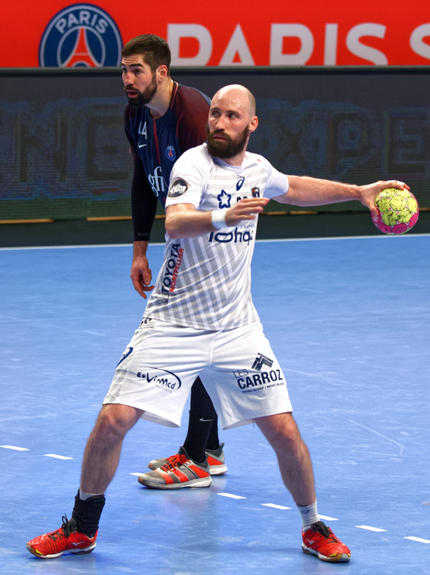 Zwei Männer spielen Handball auf einem Platz, einer hält einen Ball, mit einem Schild im Hintergrund, auf dem "Paris Saint-Germain vs Paris Saint Germain" steht.