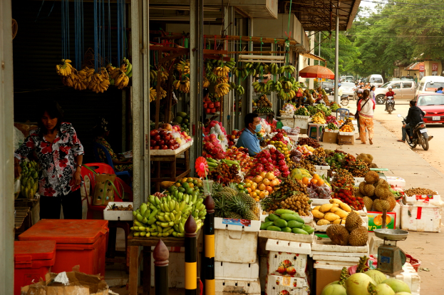 Eine belebte Straßenszene mit Bäumen, Fahrzeugen, Menschen, einige fahren Fahrzeuge, ein Obstmarkt mit verschiedenen Früchten, Behältern, einer Waage, roten Schirmen, Pfosten und Säulen.