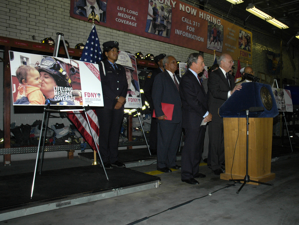 Eine Gruppe von Menschen in der Nähe eines Podiums mit Mikrofonen, Schildern auf Ständern, Flaggen und Schildern an der Wand, alles unter hellen Lichtern.