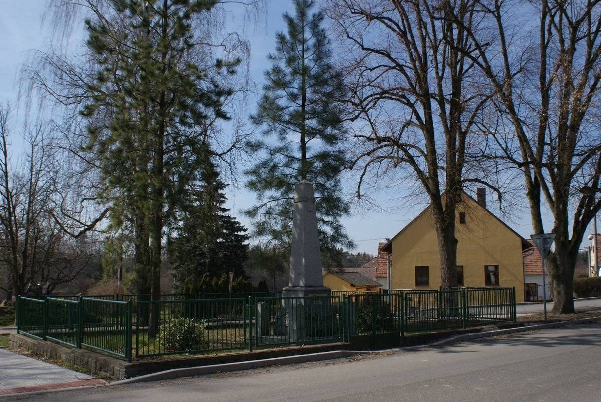 Eine Straße mit einem Holocaust-Denkmal in der Mitte, umgeben von einem Metallzaun, umgeben von Gras, Bäumen, Häusern, einem Straßenschild und einem bewölkten Himmel.