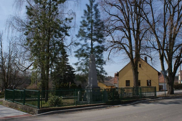 Eine Straße mit einem Holocaust-Denkmal in der Mitte, umgeben von einem Metallzaun, umgeben von Gras, Bäumen, Häusern, einem Straßenschild und einem bewölkten Himmel.