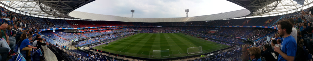 Volles Stadion mit Zuschauern bei einem Fußballspiel, einige halten Kameras, mit einem Netz auf dem Feld und sichtbaren Flutlichtmasten gegen einen klaren blauen Himmel.