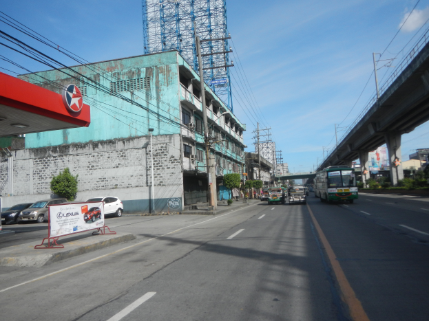Eine Stadtstraße mit Fahrzeugen, einer Tankstelle links, Gebäuden, Strommasten mit Drähten, einer Brücke rechts, Bäumen und einem bewölkten Himmel im Hintergrund.