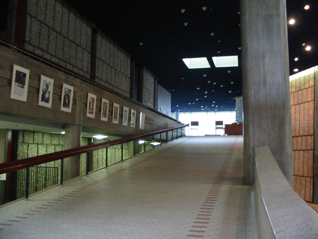 Ein Geländer auf der linken Seite mit Plakaten und Rahmen an der Wand, eine Säule auf der rechten Seite und Lampen oben.